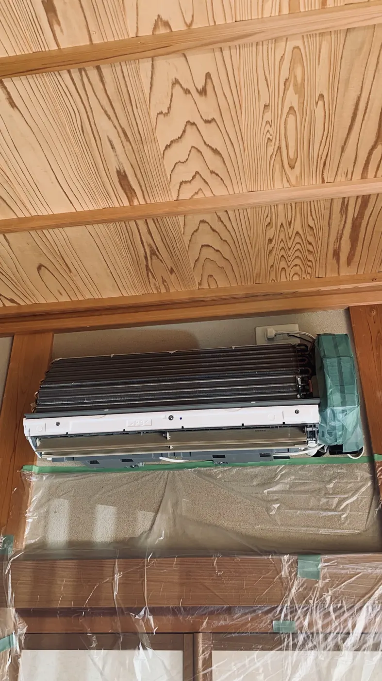 和室の壁掛けエアコンをカバーまで外し、右端の電装部を緑のシートで包み、下の壁と建具をビニールで覆っている